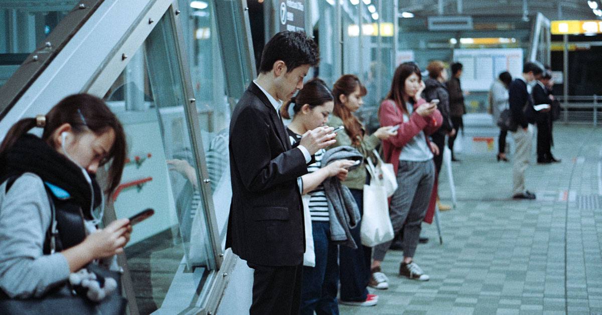 駅のホームで電車を待つ男性。スマートホンをのぞき込む